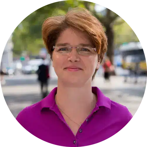 Eine Person mit kurzen braunen Haaren und Brille, die ein lila Hemd mit Kragen trägt, steht im Freien in der Nähe von Bäumen und einer unscharfen Straßenszene, möglicherweise auf dem Weg zum örtlichen Solarmarkt.