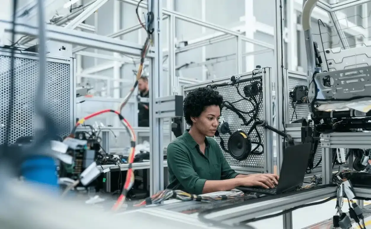 Eine Person mit kurzen lockigen Haaren arbeitet an einem Laptop in einer Hightech-Industrieumgebung, umgeben von Maschinen und elektronischen Geräten.