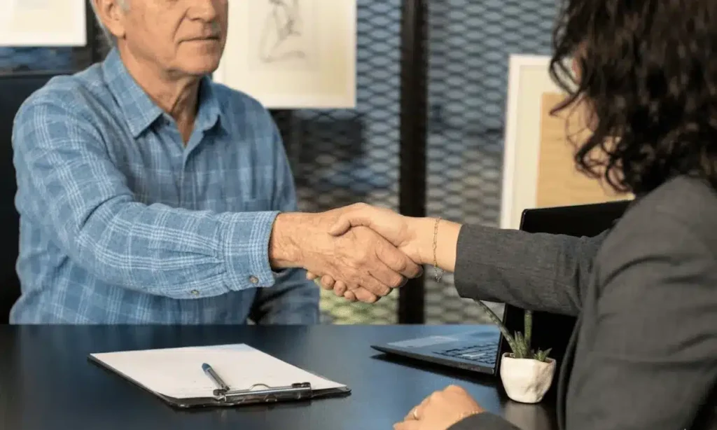 Ein älterer Mann in einem blauen Hemd und eine Frau im Anzug schütteln sich die Hände über einem Schreibtisch mit einem Klemmbrett, einem Laptop und einer kleinen Pflanze. Sie scheinen in einer professionellen Büroumgebung zu sein.