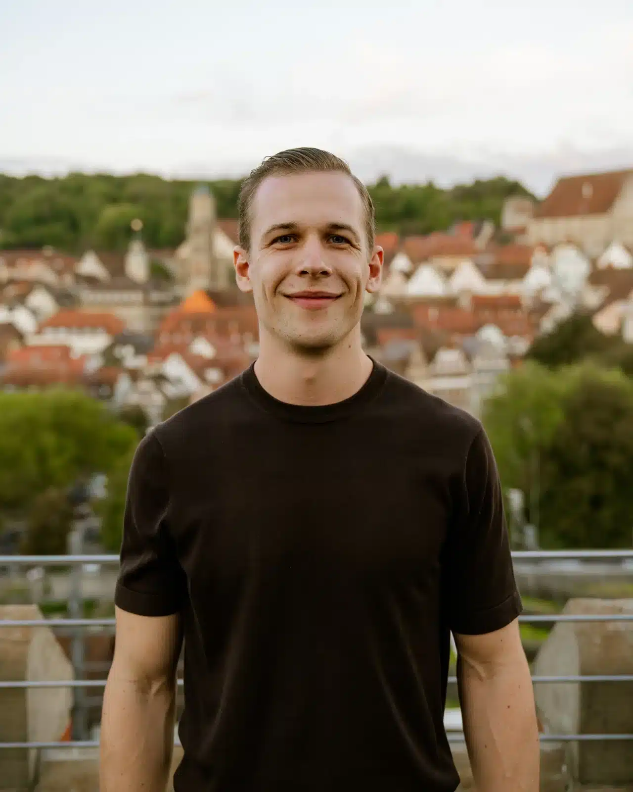 Ein Mann mit kurzen, hellbraunen Haaren und einem dunklen T-Shirt steht im Freien und lächelt in die Kamera. Hinter ihm ist eine malerische Ansicht einer Stadt mit roten Dächern, Bäumen und Hügeln zu sehen.
