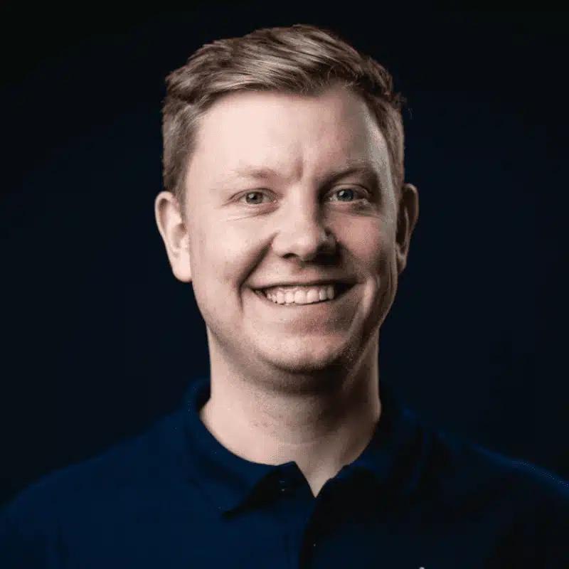 Ein lächelnder Mann mit kurzen, hellbraunen Haaren und dunkelblauem Kragenhemd steht vor einem dunklen Hintergrund und ist bereit, über die Klimaanlage als Heizlösung zu sprechen.