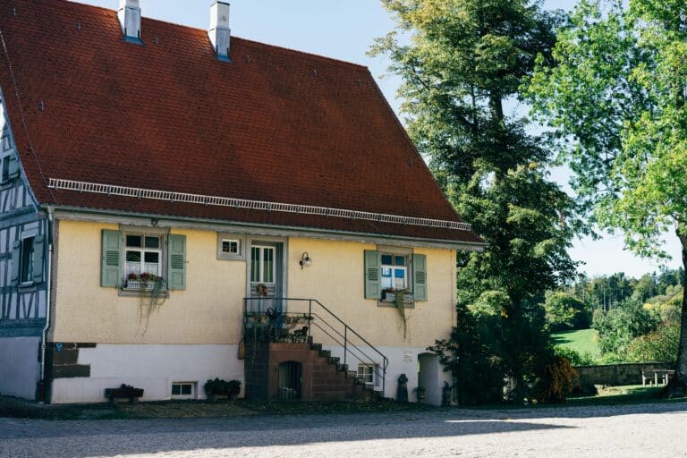 Ein charmantes Haus mit einem steilen roten Dach, hellgelben Wänden, grünen Fensterläden, Blumenkästen an den Fenstern und einer Treppe, die zur Haustür führt - jetzt mit Wärmepumpe im Altbau aufgewertet. Im Hintergrund sind Bäume und eine grüne Landschaft zu sehen.