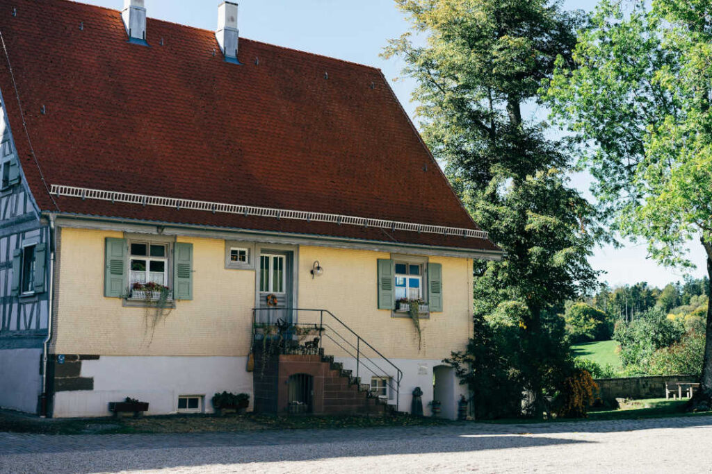 Ein charmantes Haus mit einem steilen roten Dach, hellgelben Wänden, grünen Fensterläden und Blumenkästen unter den Fenstern - jetzt mit einer effizienten Wärmepumpe im Altbau ausgestattet - liegt an einem sonnigen Tag inmitten von Bäumen und Grünflächen.