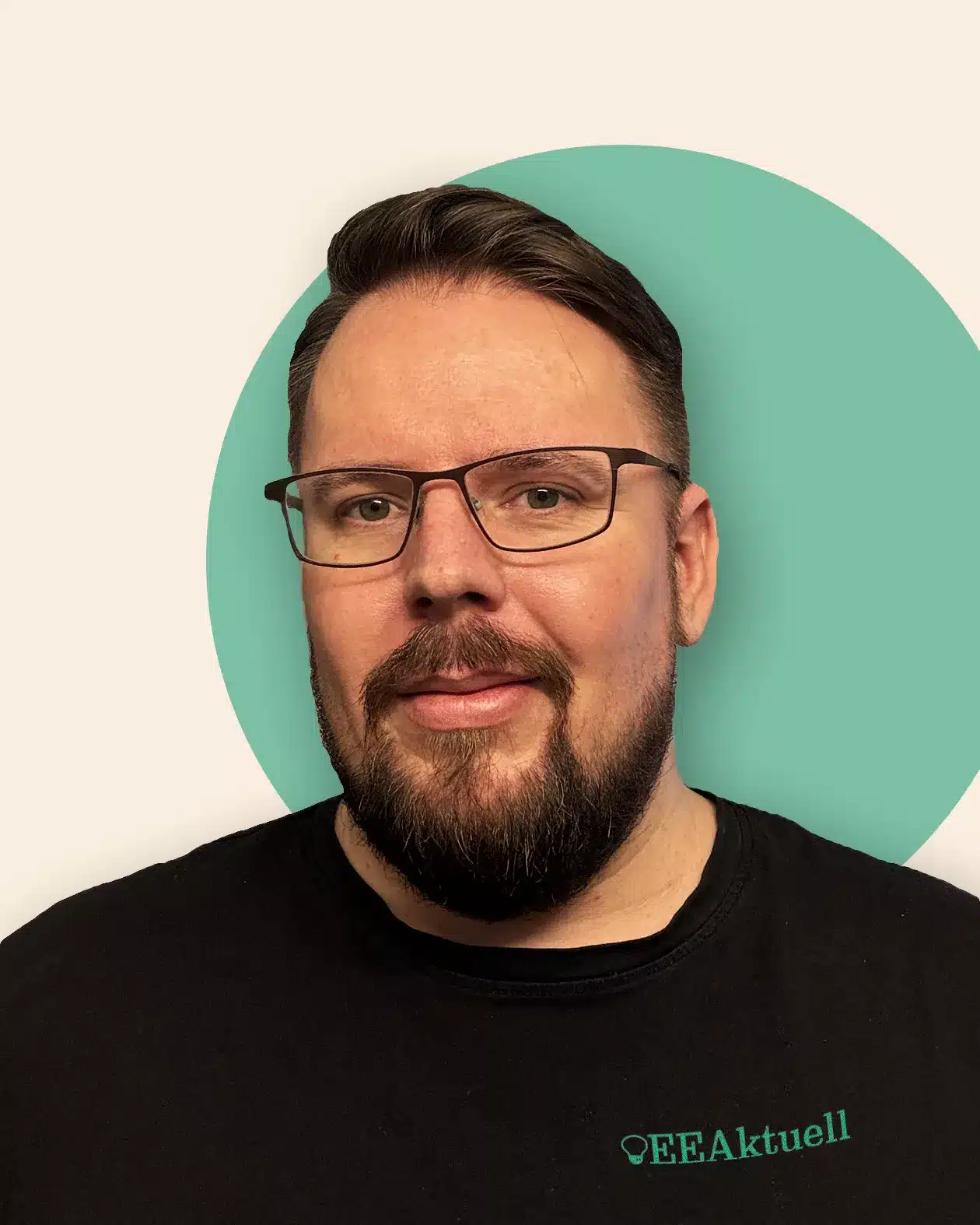 Ein bärtiger Mann mit Brille und kurzen dunklen Haaren lächelt leicht und trägt ein schwarzes "EEAktuell"-Shirt, das seine Leidenschaft für Erneuerbare Energien widerspiegelt. Der Hintergrund ist hellbeige mit einem blaugrünen Kreis hinter seinem Kopf.