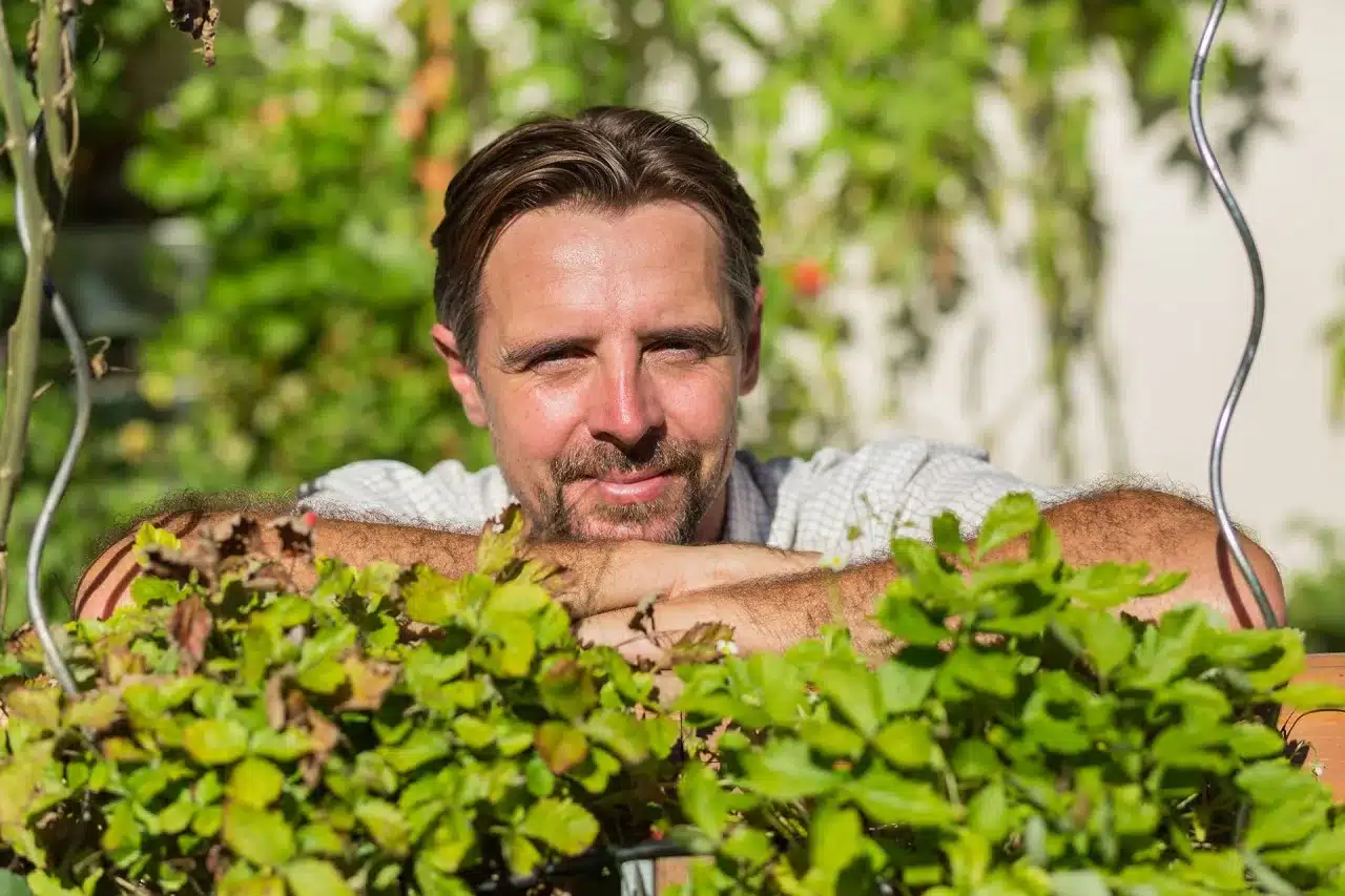 Ein Mann mit braunen Haaren und Bart lehnt lächelnd an einem Holzzaun, umgeben von üppigen grünen Pflanzen in einem sonnenbeschienenen Garten, der die Harmonie von Gebäude als Ökosystem widerspiegelt.
