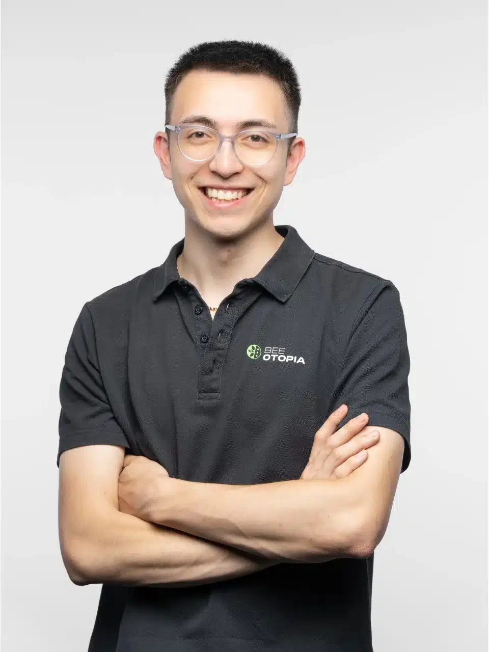 Ein junger Mann mit kurzen dunklen Haaren und Brille lächelt mit verschränkten Armen. Er trägt ein dunkles Poloshirt mit einem BEE UTOPIA-Logo und steht vor einem schlichten hellen Hintergrund - bereit, Begrünungsprojekte und Pflanzen für grüne Dächer zu unterstützen.