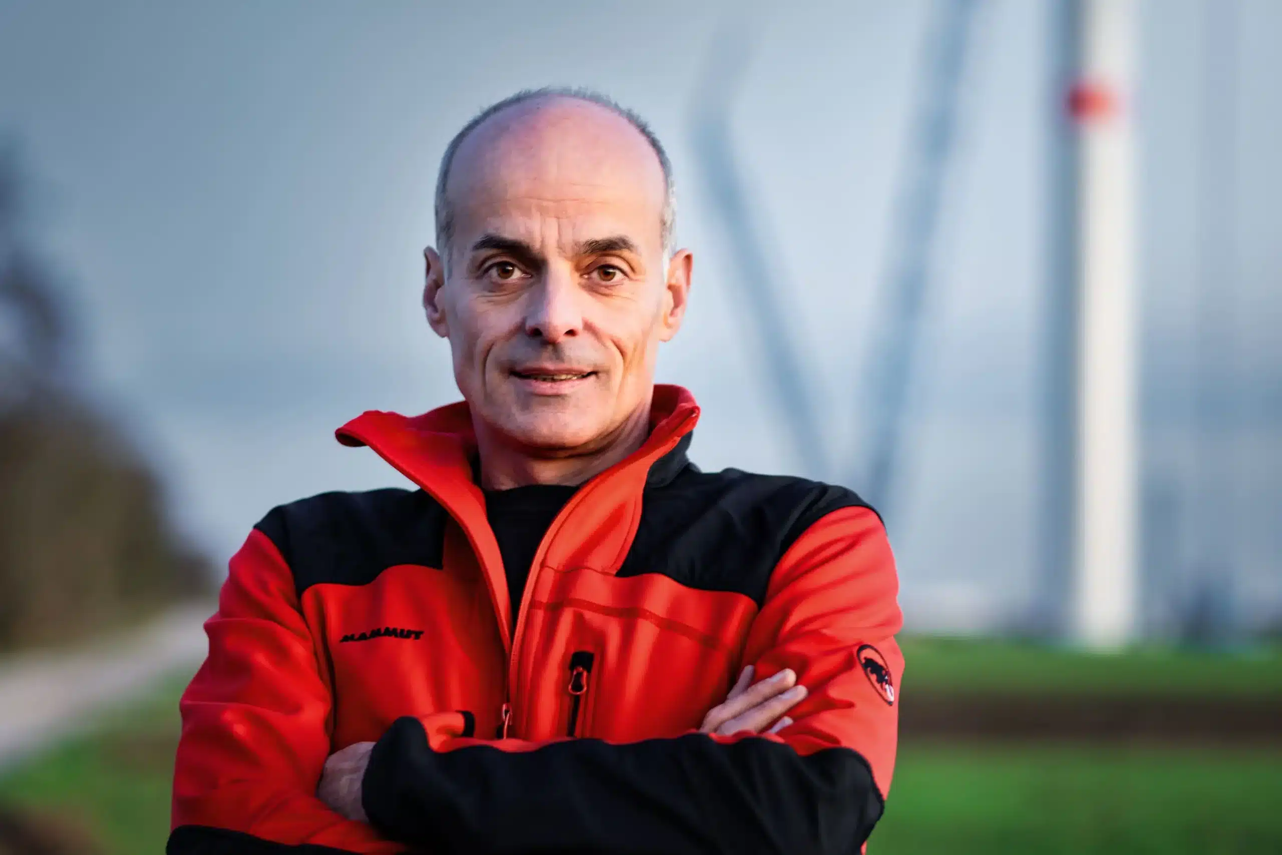 Ein Mann mittleren Alters mit kurzen Haaren, der eine rot-schwarze Jacke trägt, steht mit verschränkten Armen im Freien. Grünzeug umgibt ihn, und ein verschwommenes Windrad deutet auf die oekostrom AG Energiewende im Hintergrund hin.