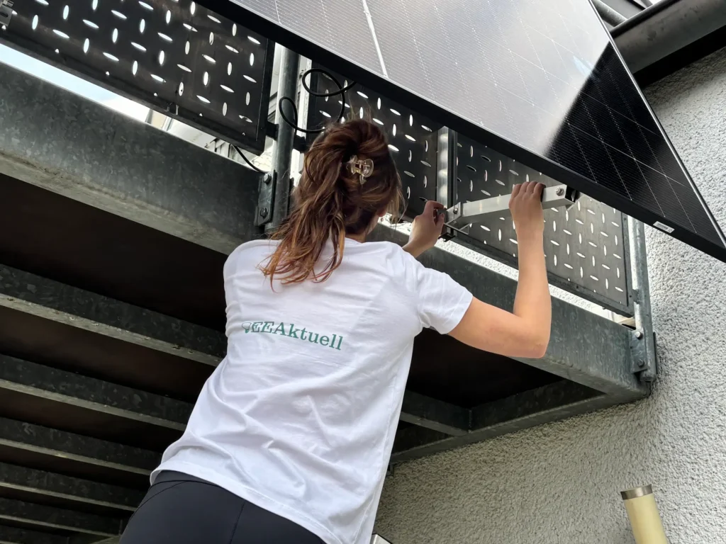 Eine Frau in weißem Hemd und schwarzer Hose steht auf einer Metallplattform und benutzt ein Werkzeug, um ein an einem perforierten Metallzaun angebrachtes Solarmodul zu installieren oder einzustellen, wobei sie sich möglicherweise von Solakon Erfahrungen leiten lässt.