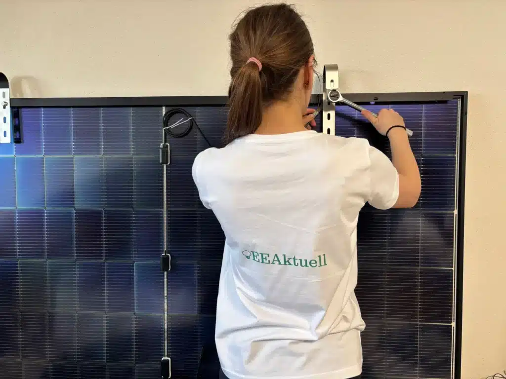 Eine Person mit braunen Haaren in einem Pferdeschwanz und einem weißen EEAktuell-T-Shirt montiert oder justiert ein Solarpanel mit einem Schraubenschlüssel an einer hellen Wand - und sammelt dabei vielleicht neue Solakon-Erfahrungen.