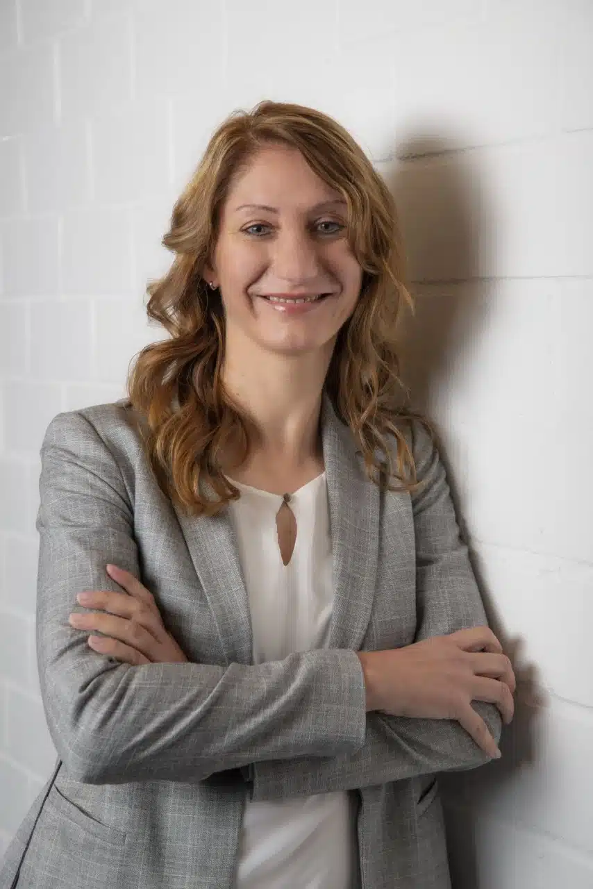 Eine Frau mit gewelltem hellbraunem Haar, hellgrauem Blazer und weißer Bluse, steht mit verschränkten Armen vor einer weißen Backsteinwand und lächelt selbstbewusst - bereit, es mit dem Alltag und dem Champion der Kreislaufwirtschaft aufzunehmen.