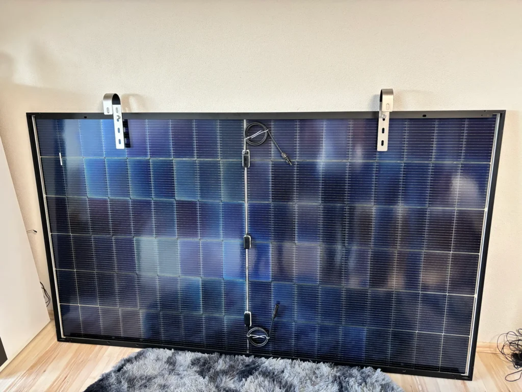 Ein großes Solarmodul, das an einer weißen Wand in einem Innenraum angebracht ist, mit zwei Metallhalterungen an der Oberseite und Stromkabeln, die von der Rückseite herabhängen - ein Beispiel für den Aufbau, der häufig in Solakon Erfahrungen zu sehen ist. Das Panel steht auf einem hellen Holzboden neben einem grauen Teppich.