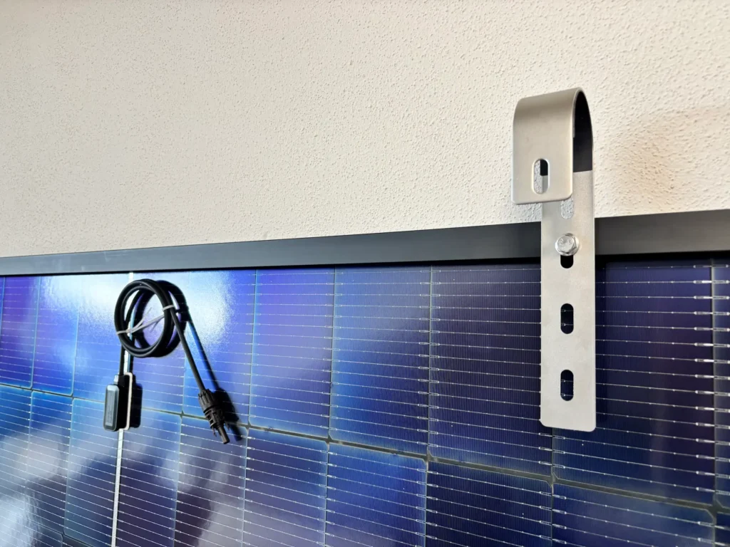 Eine Nahaufnahme eines Solarmoduls, das mit einer Metallhalterung an einer Wand befestigt ist. Ein elektrisches Kabel mit Steckern ist aufgerollt und hängt vom Rand herab. Dies spiegelt das praktische Design wider, das in vielen Solakon Erfahrungen zu sehen ist.