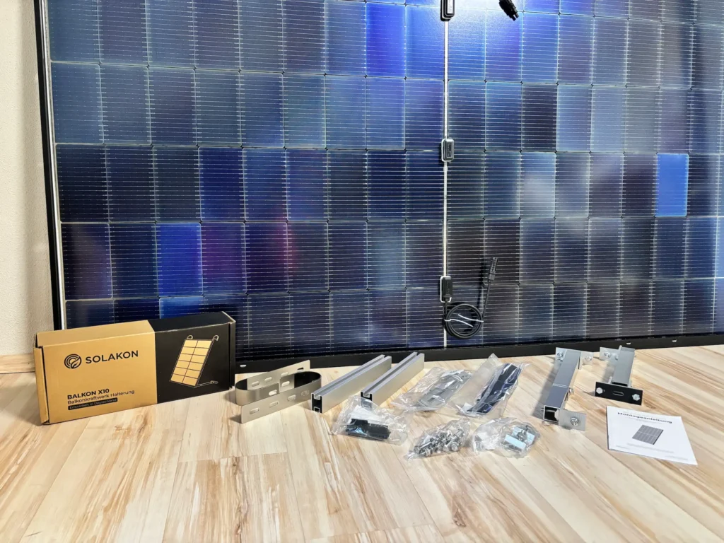 Ein Solarmodul steht auf einem Holzboden, vor dem Montagehalterungen, Schrauben, Beschläge, Anleitungen und eine Schachtel mit der Marke Solakon liegen - perfekt für alle, die bei der Montage echte Solakon-Erfahrungen sammeln wollen.