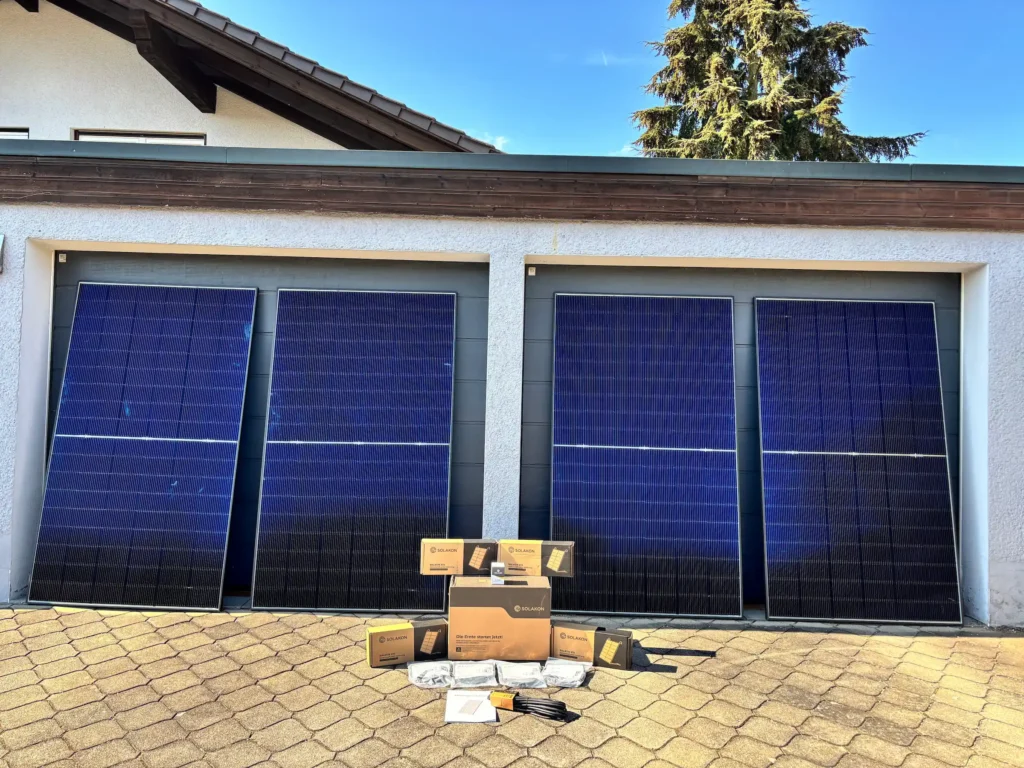 Vier große blaue Solarmodule sind gegen ein weißes Garagentor gelehnt. Davor sind mehrere Pappkartons, Kabel und Bedienungsanleitungen auf einer gepflasterten Auffahrt angeordnet - eine typische Einrichtung von Solakon Erfahrungen. Darüber erscheinen ein Baum und eine Dachlinie.