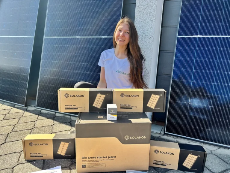 Eine lächelnde Frau in einem weißen T-Shirt sitzt im Freien, vor sich mehrere Boxen der Marke SOLAKON. Im Hintergrund sind große Solarpaneele zu sehen, die auf eine Umgebung hinweisen, in der Solakon-Erfahrungen weitergegeben werden.