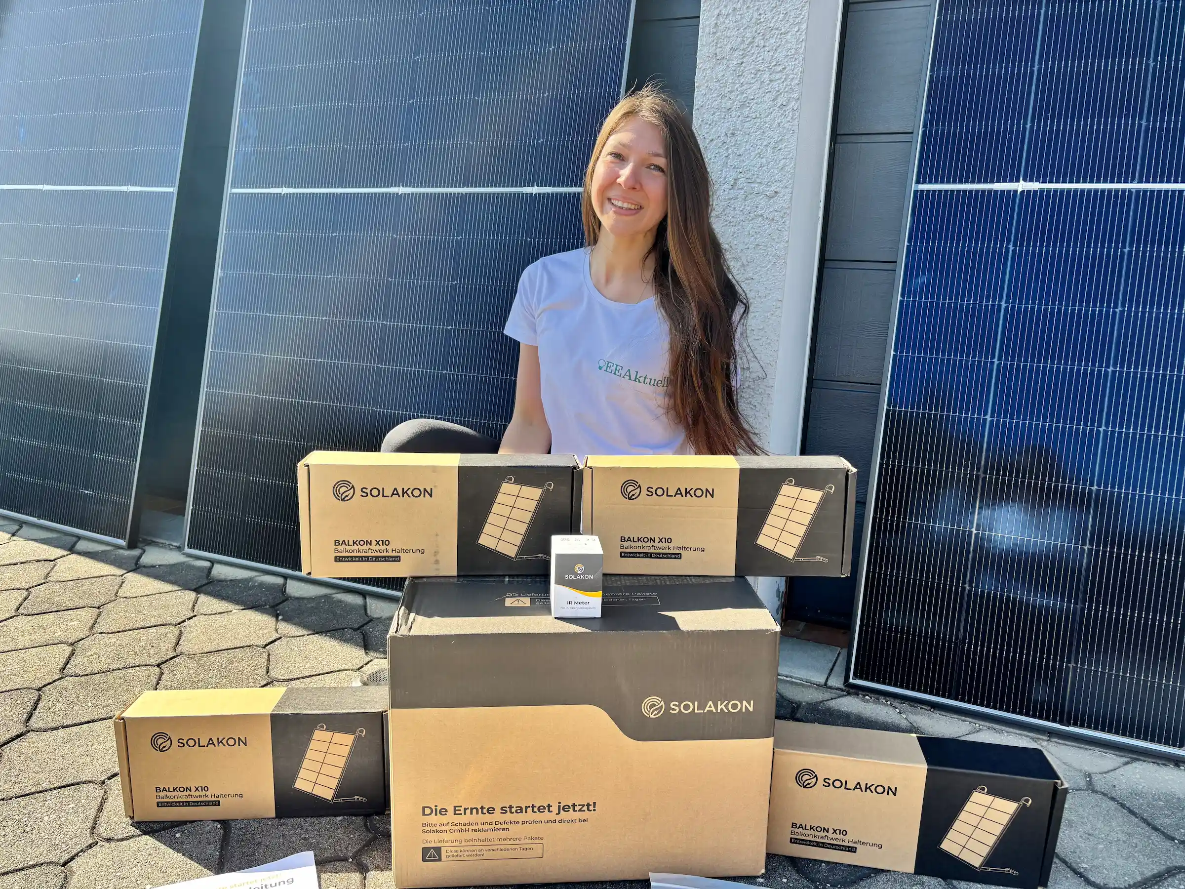 Eine lächelnde Frau in einem weißen T-Shirt sitzt im Freien, vor sich mehrere Boxen der Marke SOLAKON. Im Hintergrund sind große Solarpaneele zu sehen, die auf eine Umgebung hinweisen, in der Solakon-Erfahrungen weitergegeben werden.