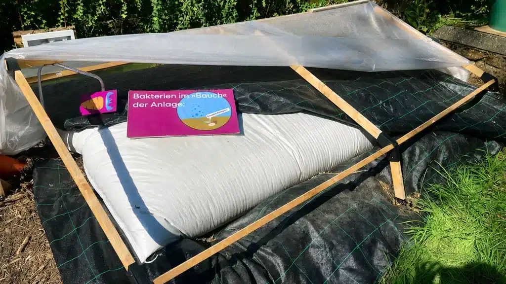 Ein überdachtes Gartenbeet mit einem Holzrahmen und einer Plastikfolie, auf dem ein Schild in deutscher Sprache mit der Aufschrift "Bakterien im Bauch der Anlage" und einer Abbildung von Bakterien zu sehen ist - perfekt als Mini-Biogasanlage für den deutschen Markt. Gras und Grünzeug umgeben die Anlage.
