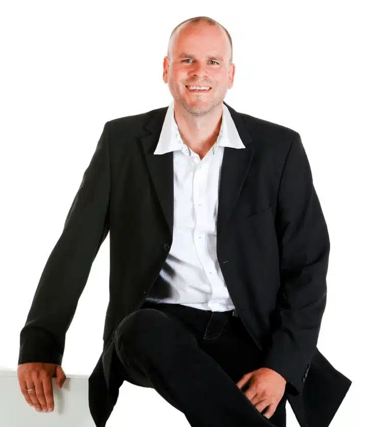 Ein lächelnder Mann mit kurzem Haar, gekleidet in einen schwarzen Blazer, ein weißes Hemd und eine schwarze Hose, sitzt lässig mit einer Hand auf einer weißen Fläche vor einem schlichten weißen Hintergrund und verkörpert den innovativen Geist von Windmarkt und neue Strukturen.