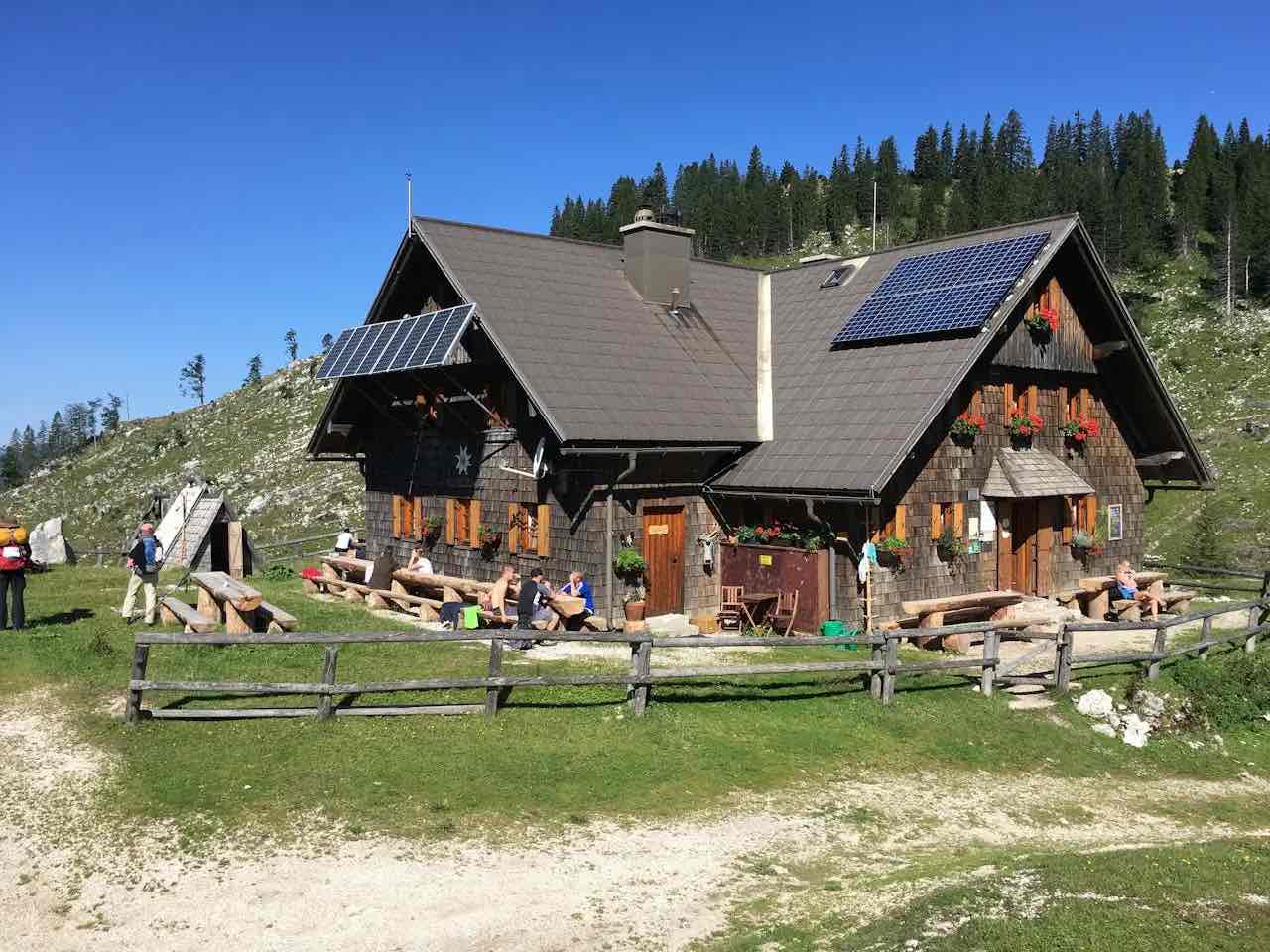 Eine rustikale Berghütte mit Sonnenkollektoren auf dem Dach ist ein Beispiel für cleveres Energiemanagement im Eigenheim, umgeben von Bänken und Holzzäunen in einer grasbewachsenen Alpenlandschaft unter einem strahlend blauen Himmel. Mehrere Menschen entspannen sich in der Nähe.