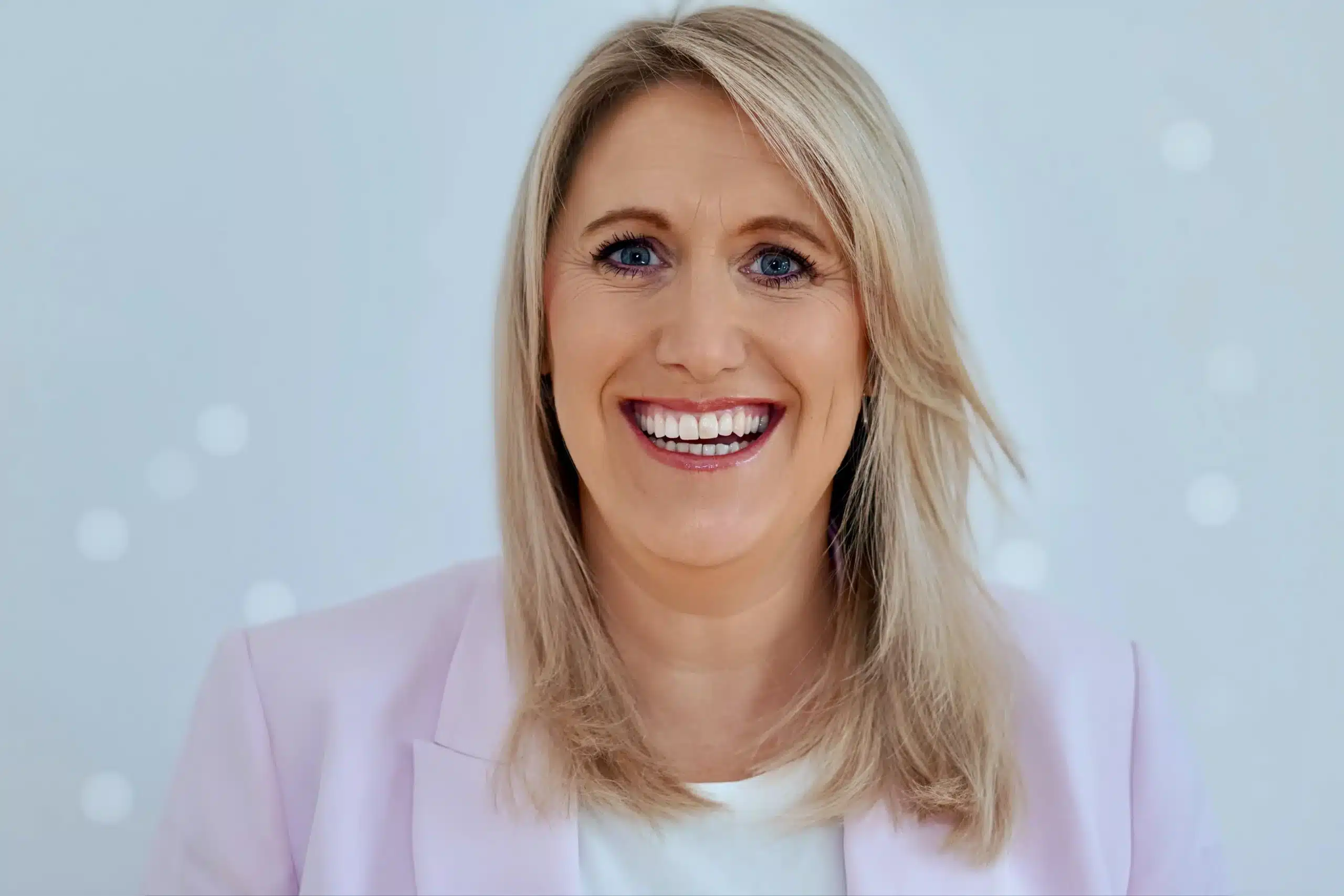 Eine Frau mit glattem blondem Haar und blauen Augen lächelt breit. Sie trägt einen hellrosa Blazer über einem weißen Oberteil vor einem hellblauen Hintergrund – und verkörpert damit perfekt das moderne Energiemanagement im Eigenheim.