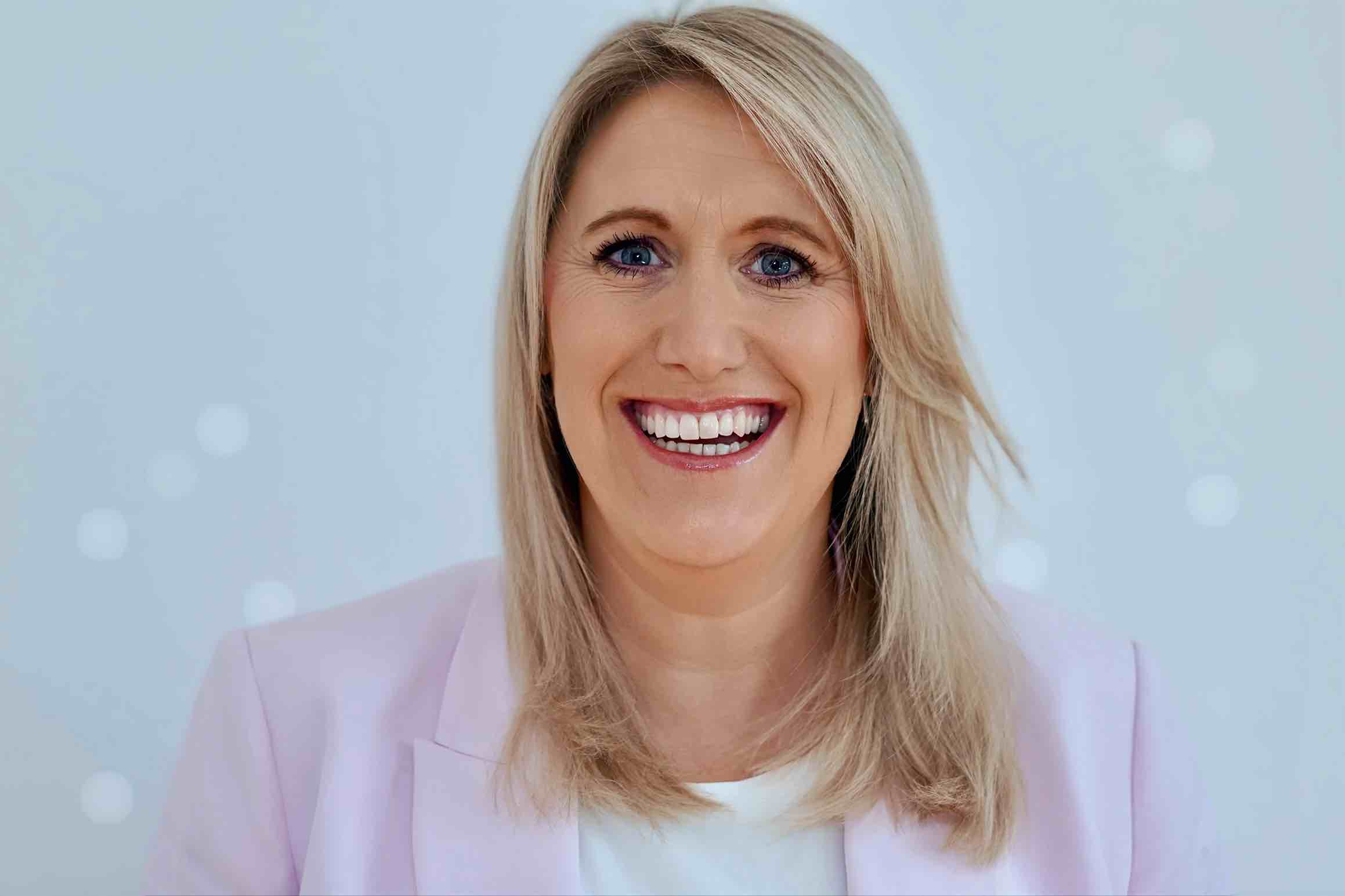 Eine Frau mit blondem Haar und blauen Augen lächelt breit. Sie trägt einen hellrosa Blazer über einem weißen Oberteil und strahlt vor einem hellen, leicht verschwommenen Hintergrund Zuversicht aus - vielleicht nachdem sie Energiemanagement im Eigenheim gemeistert hat.