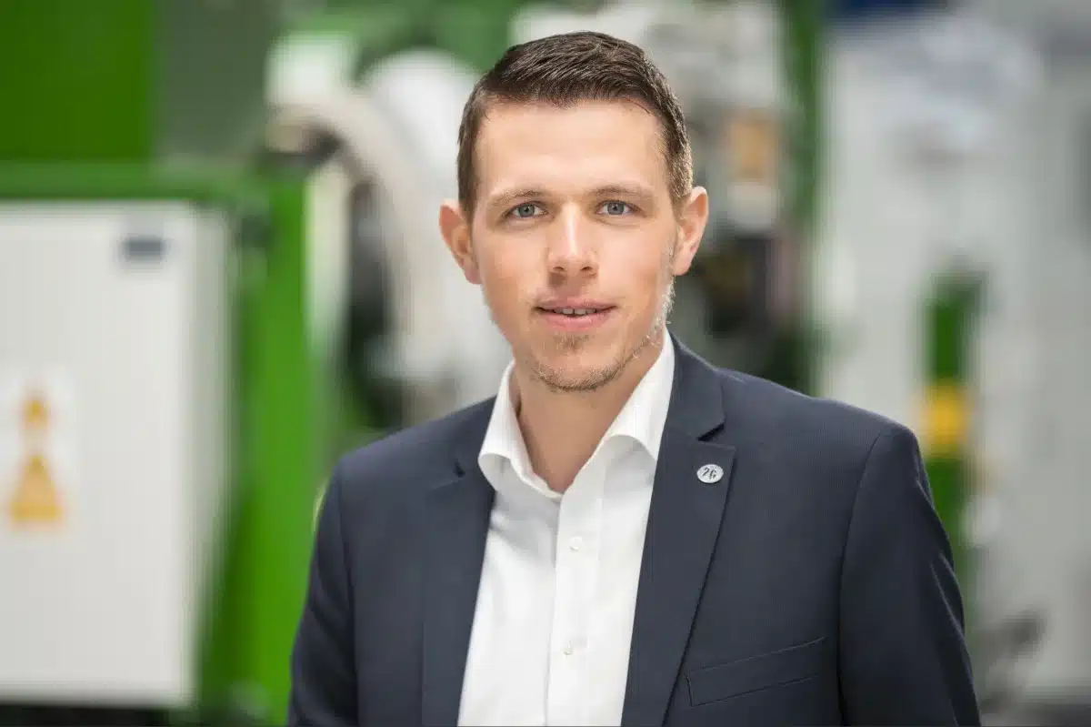Ein Mann mit kurzen braunen Haaren, der ein dunkles Sakko und ein weißes Hemd trägt, steht in einem Innenraum und blickt in die Kamera, im Hintergrund sind verschwommene grüne und weiße Blockheizkraftwerke zu sehen.