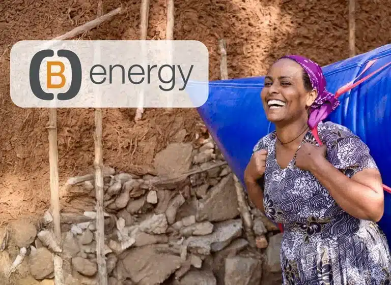Eine lächelnde Frau in einem gemusterten Kleid trägt einen großen blauen Behälter auf dem Rücken vor einer Lehmwand. Das B Energy-Logo und der Schriftzug "(B)Energy Erfahrungen" erscheinen in der oberen linken Ecke des Bildes.