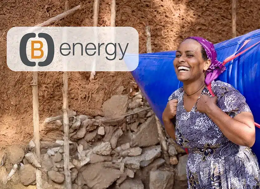 Eine lächelnde Frau in einem gemusterten Kleid trägt einen großen blauen Behälter auf dem Rücken vor einer Lehmwand. Das B Energy-Logo und der Schriftzug "(B)Energy Erfahrungen" erscheinen in der oberen linken Ecke des Bildes.
