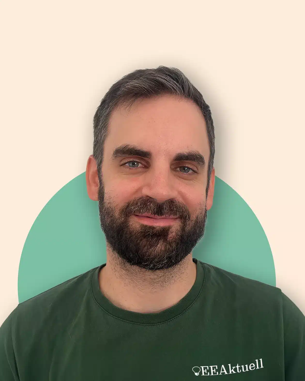 Ein Mann mit kurzen dunklen Haaren und Bart, der ein grünes „EEAktuell“-T-Shirt trägt, steht vor einem beigen Hintergrund, der durch einen grünen Halbkreis hervorgehoben wird. Er lächelt sanft und repräsentiert damit die Marke „EEAktuell“.