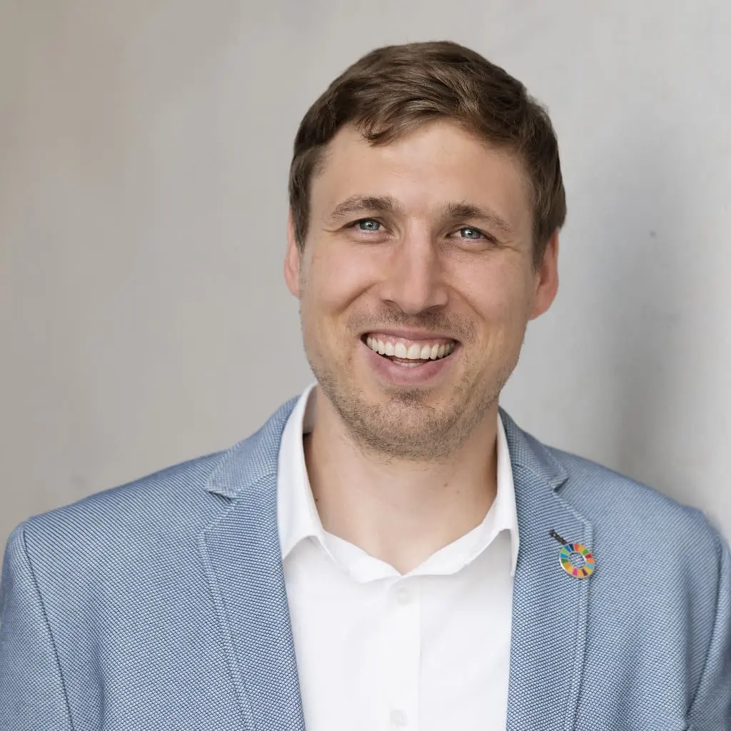 Ein lächelnder Mann mit kurzen braunen Haaren, der einen hellblauen Blazer, ein weißes Hemd und eine bunte Anstecknadel am Revers trägt, steht vor einem schlichten hellen Hintergrund – bereit, die Energiewende im Betrieb voranzutreiben.
