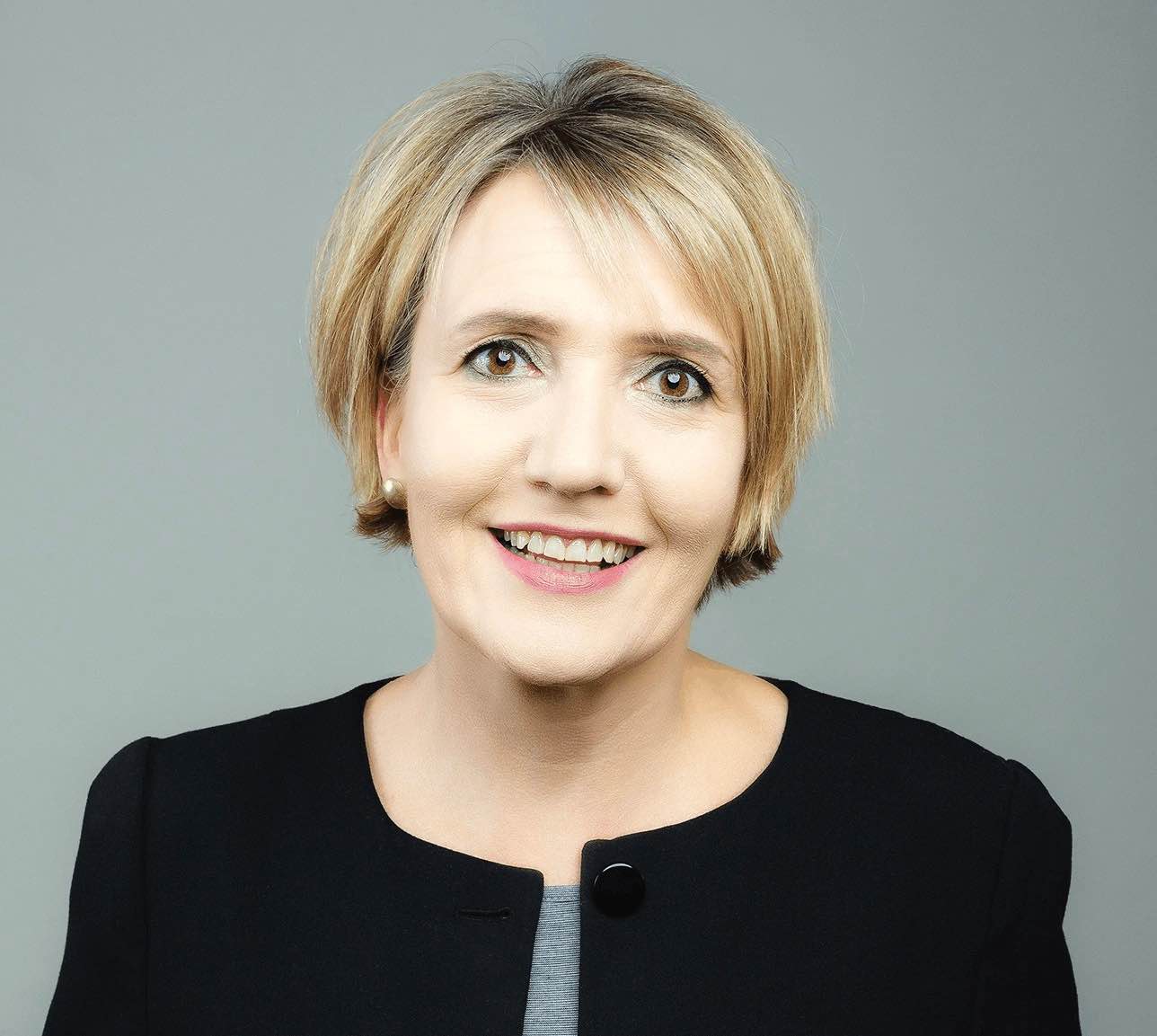 Eine Frau mit kurzen blonden Haaren, die einen schwarzen Blazer über einem grauen Oberteil trägt, lächelt vor einem schlichten grauen Hintergrund in die Kamera – ein selbstbewusster und zugänglicher Ausdruck, der an Simone Peter erinnert, während sie sich für die Energiewende 2026 einsetzt.