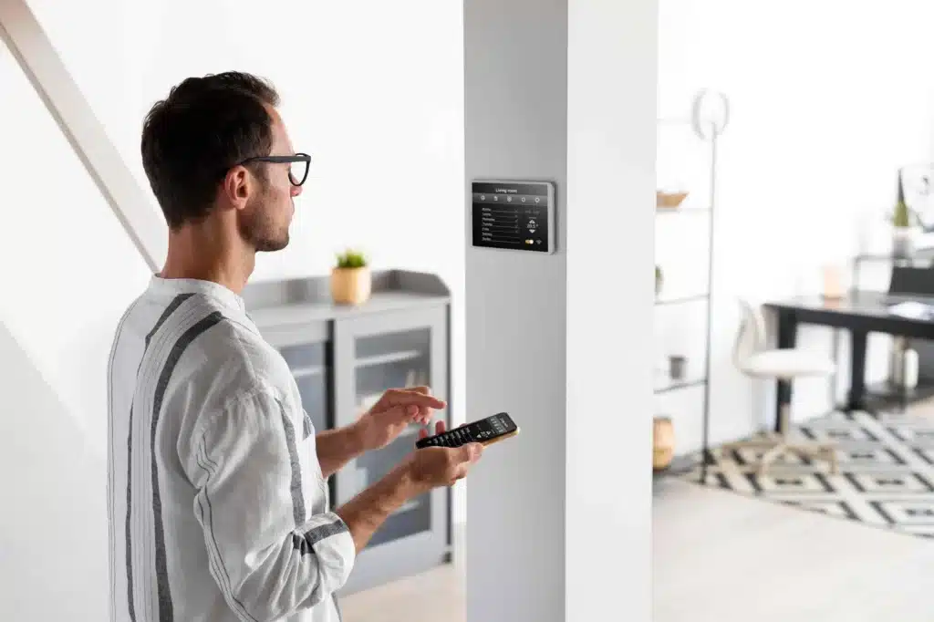 Ein Mann, der ein Smartphone und eine Fernbedienung in der Hand hält, steht in einem modernen Wohnraum neben einem an der Wand montierten Smart-Home-Bedienfeld und erkundet Möglichkeiten, die Nachtspeicherheizung durch die Einbindung von erneuerbarem Strom zu modernisieren.