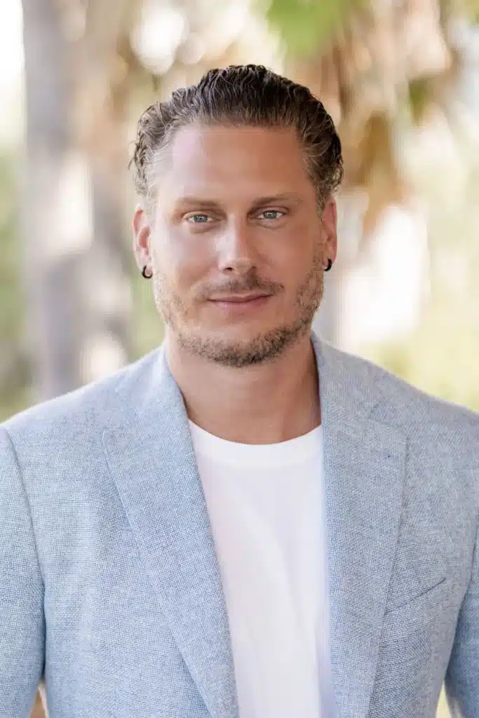 Ein Mann mit kurzen, nach hinten geglätteten Haaren und leichtem Bartwuchs trägt einen hellblauen Blazer und ein weißes Hemd und steht im Freien an einem erstklassigen Standort, im Hintergrund sind verschwommene Grünflächen, Palmblätter und Andeutungen auf Investitionsmöglichkeiten im Bereich Großbatteriespeicher zu sehen.