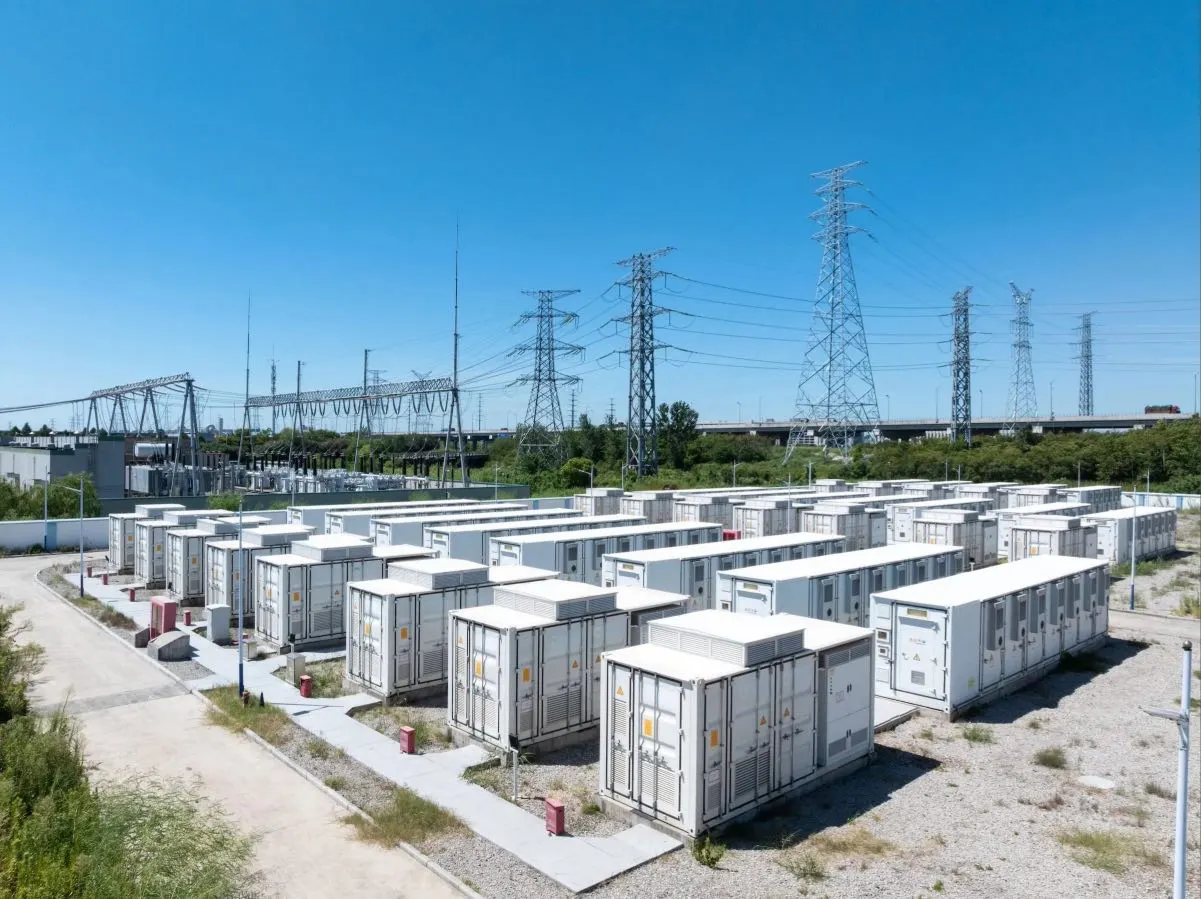Reihen großer weißer Batteriespeicher, sogenannte Großbatteriespeicher, sind an diesem strategisch wichtigen Standort unter strahlend blauem Himmel im Freien in der Nähe von Stromleitungen und Strommasten angeordnet, umgeben von Schotterwegen und etwas Vegetation.