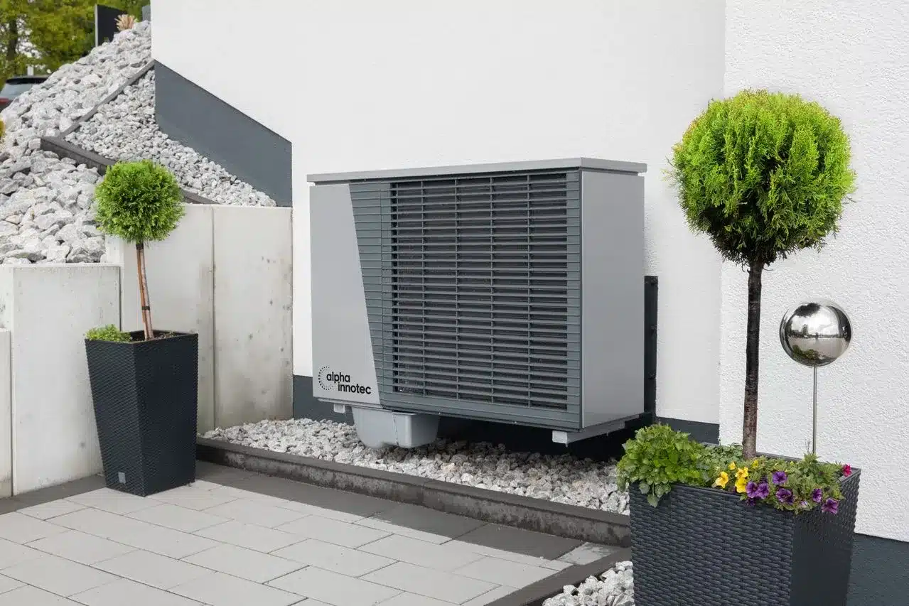Eine moderne Wärmepumpenanlage, die außerhalb eines Gebäudes installiert ist und von Zierkies, Topfpflanzen und einer weißen Mauer in einem gepflegten Außenbereich umgeben ist – ein hervorragendes Beispiel dafür, wie man mit einer Wärmepumpe die Gasheizung ersetzen und so energieeffiziente Heizlösungen realisieren kann.