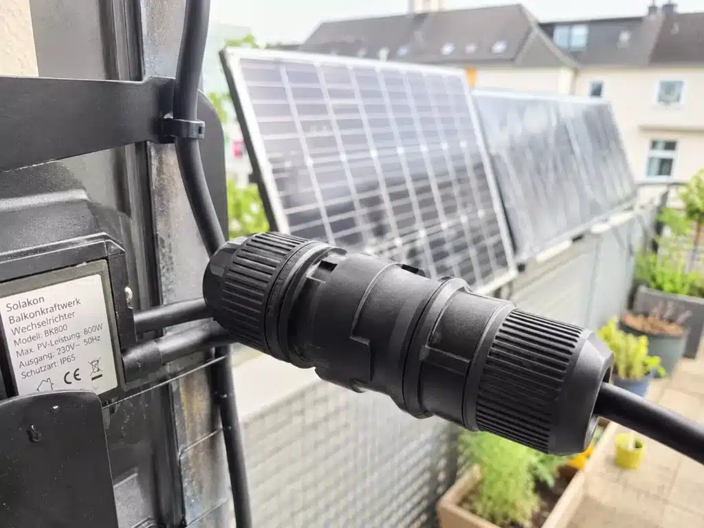 Nahaufnahme eines schwarzen Steckverbinders, der an einer Solaranlage auf einem Balkon angebracht ist und über eine sichere Wieland-Steckdose verfügt. Im Hintergrund sind mehrere Solarmodule und Pflanzen zu sehen.