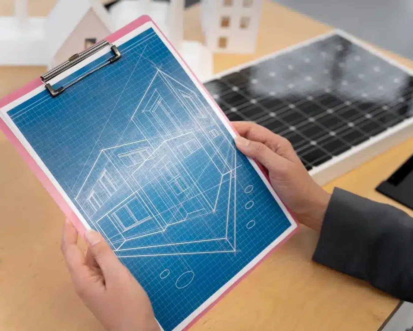 Eine Person hält ein Klemmbrett mit der Bauzeichnung eines modernen Hauses. Im Hintergrund sind auf dem Schreibtisch ein Solarpanel und architektonische Gebäudemodelle zu sehen.