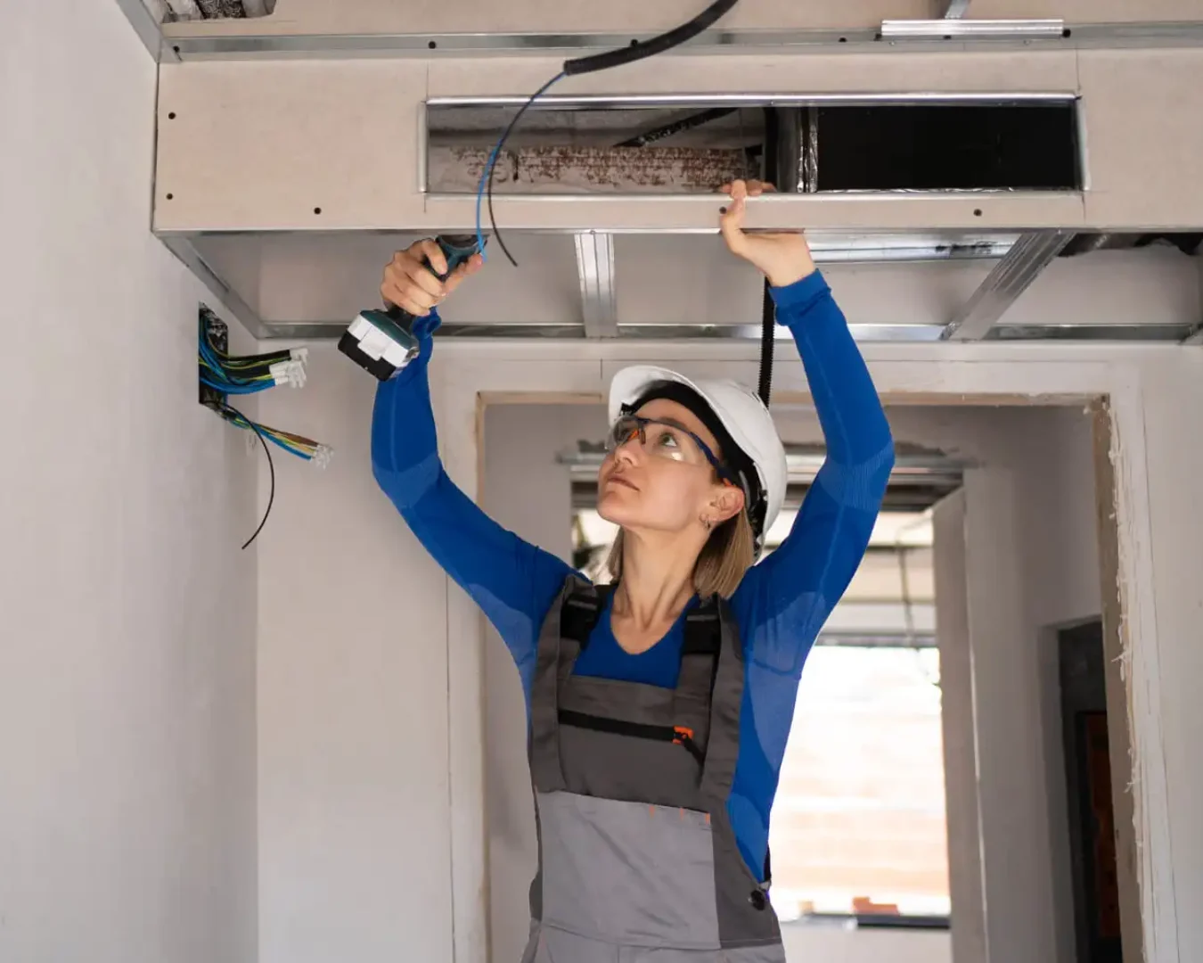Eine Frau, die einen Schutzhelm und eine Schutzbrille trägt, arbeitet mit einer Bohrmaschine an elektrischen Leitungen in einer Decke, um energieeffiziente Lüftungssysteme in einem teilweise gebauten oder renovierten Raum vorzubereiten.