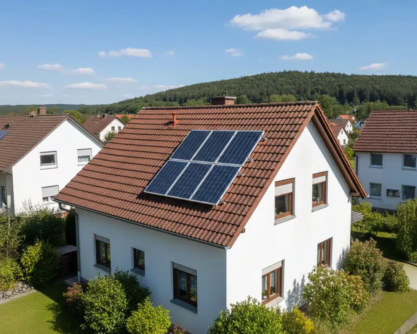 Ein weißes Haus mit rotem Ziegeldach und Sonnenkollektoren demonstriert, wie viel Strom eine 10 kWp Photovoltaikanlage am Tag produzieren kann. Es ist umgeben von grünen Büschen und Bäumen, von ähnlichen Häusern und einem bewaldeten Hügel unter einem blauen, wolkenverhangenen Himmel.