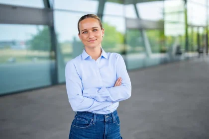 Eine Frau mit hellbraunem, zurückgebundenem Haar, hellblauem Button-up-Hemd und Jeans steht mit verschränkten Armen und einem selbstbewussten Lächeln vor einem modernen Glasgebäude, das Nachhaltigkeit in Unternehmen widerspiegelt.