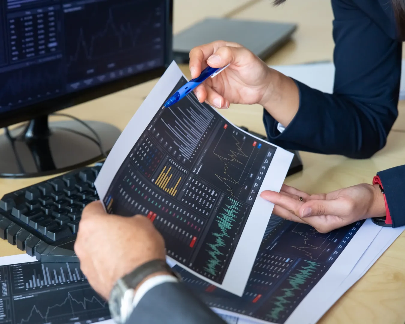 Zwei Personen in Geschäftskleidung prüfen an einem Schreibtisch gedruckte Diagramme und Grafiken mit Finanzdaten, daneben ein Monitor, auf dem ETF Sparplan Vergleich und ähnliche Finanzinformationen angezeigt werden.