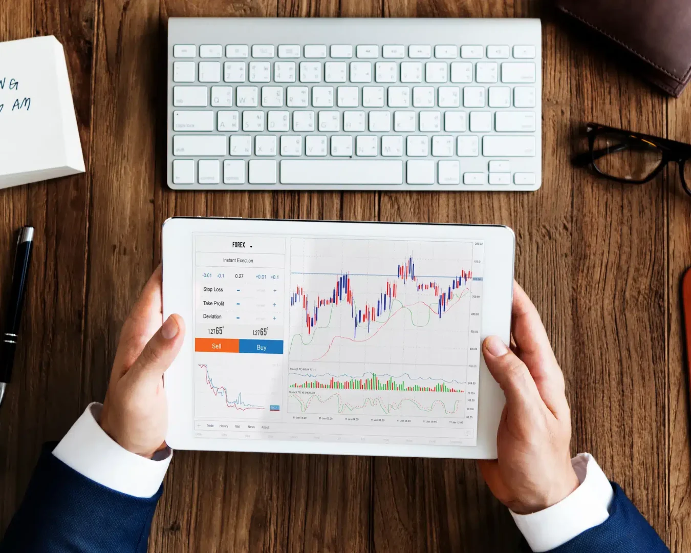 Eine Person im Anzug hält ein Tablet, auf dem ein Aktienchart mit Kauf- und Verkaufsoptionen angezeigt wird, neben einer drahtlosen Tastatur und einem Notizblock mit MEETING @ 9.30 AM - sie recherchiert die besten ETF-Sparplan-Strategien.