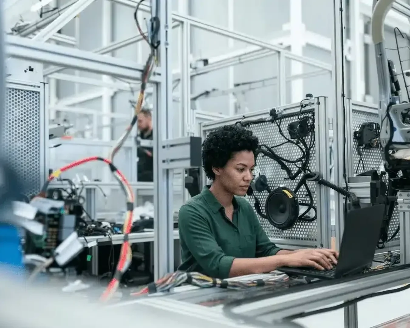 Eine Person mit kurzen lockigen Haaren arbeitet an einem Laptop in einer Hightech-Industrieumgebung, umgeben von Maschinen und elektronischen Geräten.