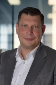 Marian Klemm, ein Mann mit kurzen braunen Haaren, der eine graue Anzugsjacke und ein weißes Hemd trägt, lächelt leicht und schaut in die Kamera. Der Hintergrund ist eine Büroumgebung mit Glaswänden und unscharfem Licht.