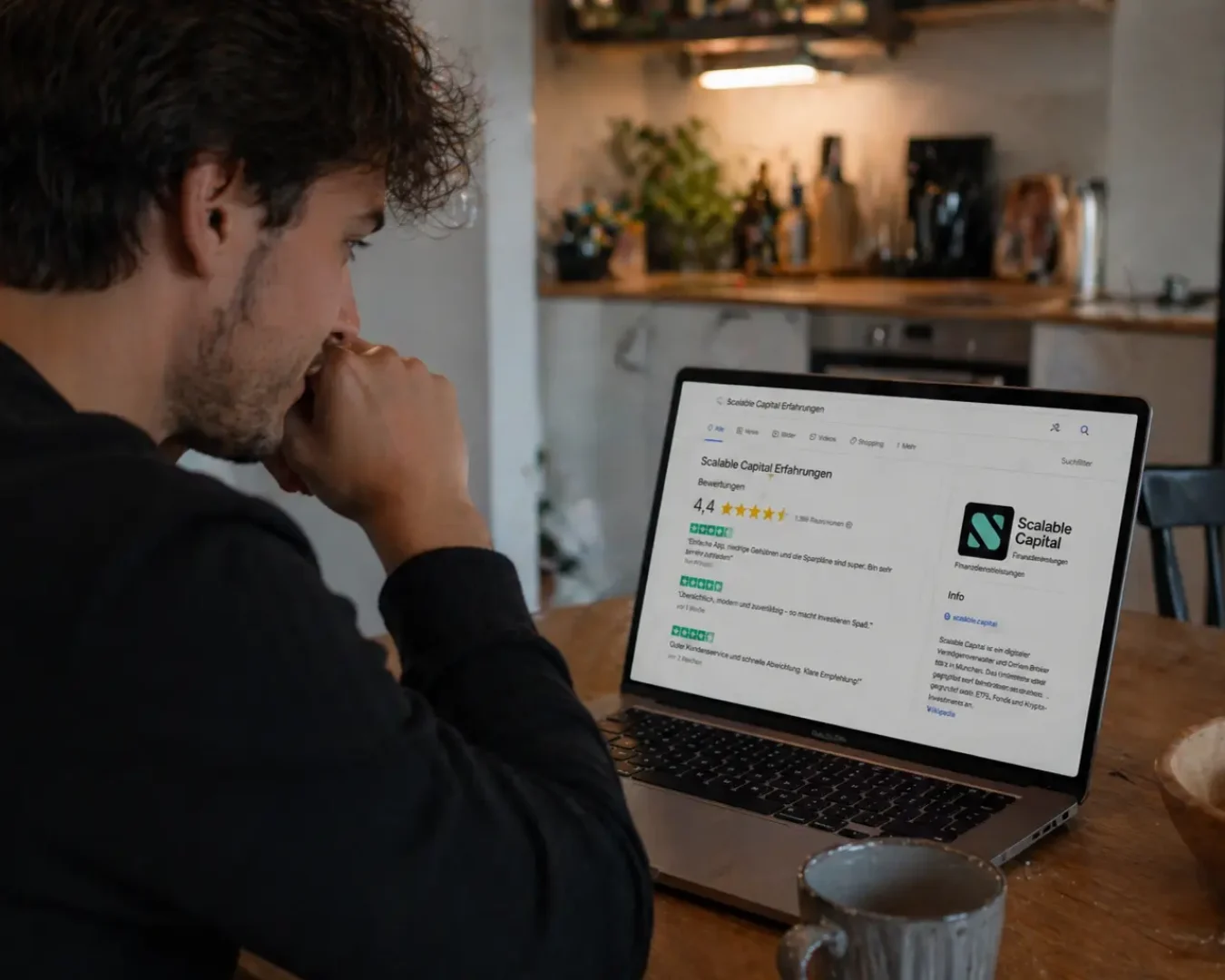 Ein Mann sitzt an einem Tisch in einer Küche und schaut nachdenklich auf einen Laptop-Bildschirm, auf dem Scalable Capital Erfahrungen und Informationen anzeigt. Eine Tasse und eine Schüssel stehen auf dem Tisch neben ihm.