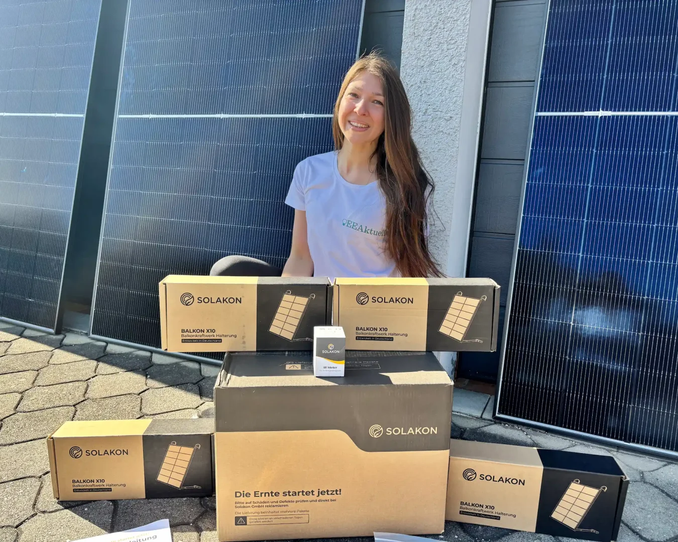 Eine lächelnde Frau in einem weißen T-Shirt sitzt im Freien, vor sich mehrere Boxen der Marke SOLAKON. Im Hintergrund sind große Solarpaneele zu sehen, die auf eine Umgebung hinweisen, in der Solakon-Erfahrungen weitergegeben werden.