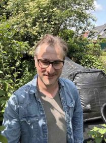 Ein Mann mit Brille und Jeanshemd steht draußen in einem üppigen, grünen Garten, umgeben von Bäumen und Pflanzen, im Hintergrund ein mit einer schwarzen Plane bedecktes Bioenergieobjekt.