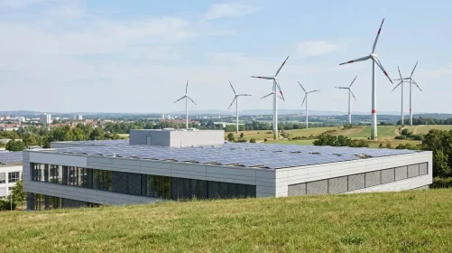 Ein modernes Gebäude mit Sonnenkollektoren auf dem Dach steht auf einer Wiese, im Hintergrund sind unter einem teilweise bewölkten Himmel mehrere Windkraftanlagen zu sehen. Am Horizont ist eine ferne Stadt zu erkennen, was an die Themen erinnert, die im jüngsten ZSW-Energiewende-Interview über nachhaltige Energielösungen und zukünftige Stadtlandschaften diskutiert wurden.