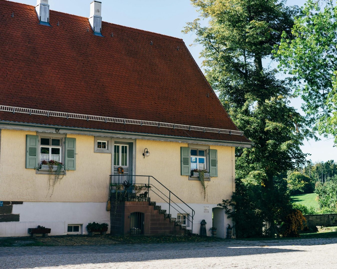 Ein charmantes Haus mit einem steilen roten Dach, hellgelben Wänden, grünen Fensterläden, Blumenkästen an den Fenstern und einer Treppe, die zur Haustür führt - jetzt mit Wärmepumpe im Altbau aufgewertet. Im Hintergrund sind Bäume und eine grüne Landschaft zu sehen.
