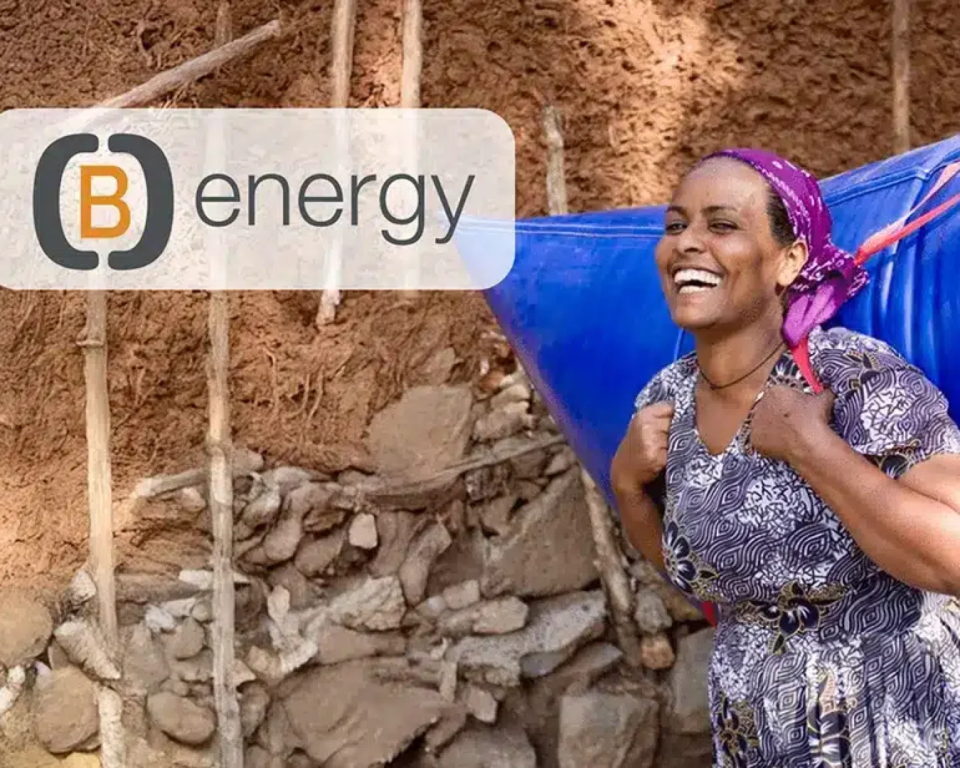 Eine lächelnde Frau in einem gemusterten Kleid trägt einen großen blauen Behälter auf dem Rücken vor einer Lehmwand. Das B Energy-Logo und der Schriftzug "(B)Energy Erfahrungen" erscheinen in der oberen linken Ecke des Bildes.